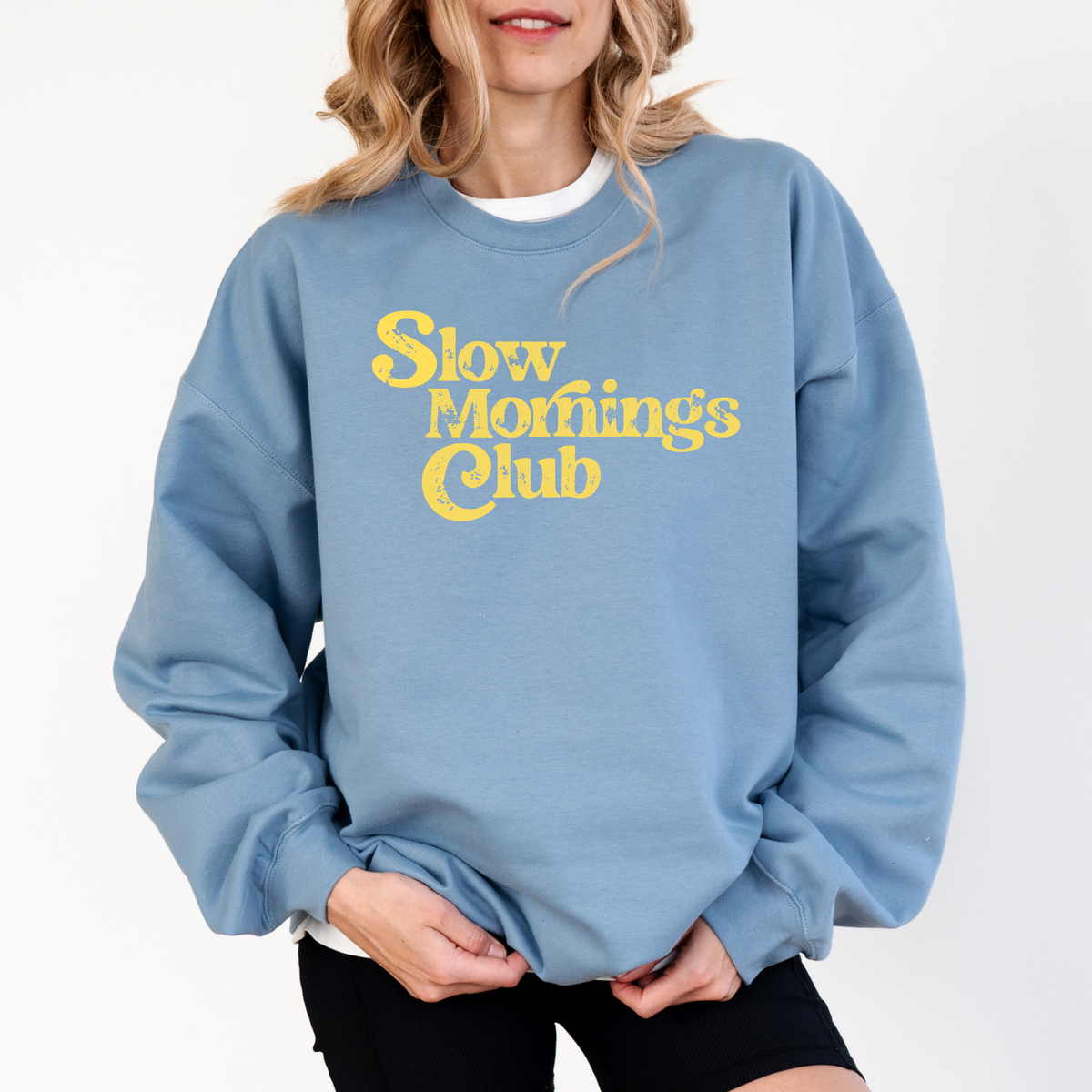 A woman wearing a stone blue sweatshirt with 'Slow mornings club' text in yellow on a white background