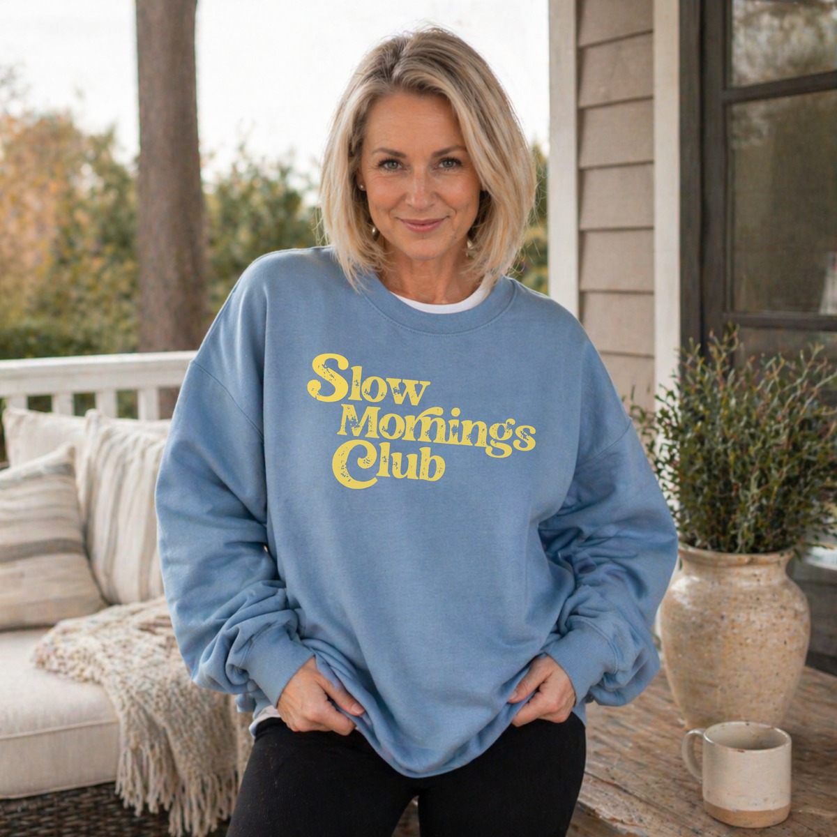 Woman wearing a blue sweatshirt with 'Slow Mornings Club' text on a porch.