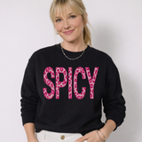 Person wearing a black sweater with 'SPICY' in pink leopard print letters on a plain background