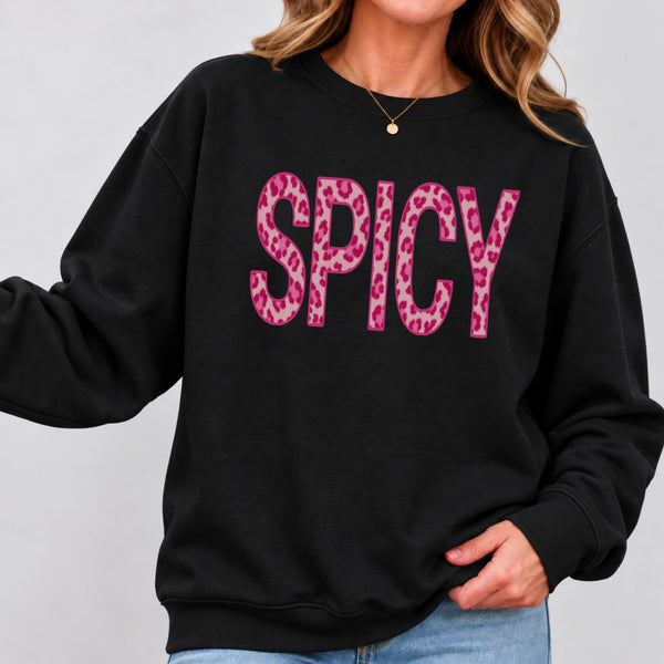 A woman wearing a black crewneck sweatshirt with the word 'SPICY' printed across the chest in bold pink letters filled with a pink leopard print pattern, paired with light blue jeans against a light gray background.