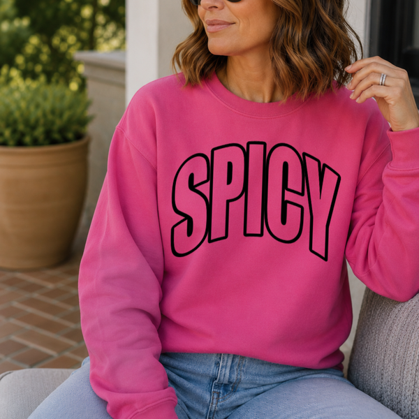 A woman wearing a bright pink crewneck sweatshirt with the word 'SPICY' printed across the chest in large black outlined letters, sitting on a gray chair.