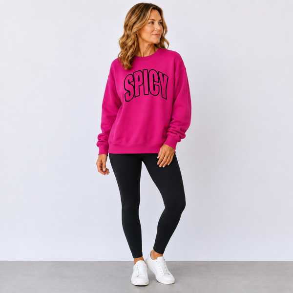 A woman modeling a bright pink crewneck sweatshirt with 'SPICY' printed in black arched letters across the chest, paired with black leggings and white sneakers, against a light gray studio background.