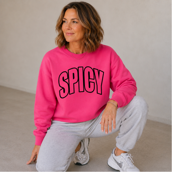 A woman crouching in a bright pink sweatshirt with the word 'SPICY' printed across the chest in black outline lettering, paired with light gray sweatpants and white sneakers.