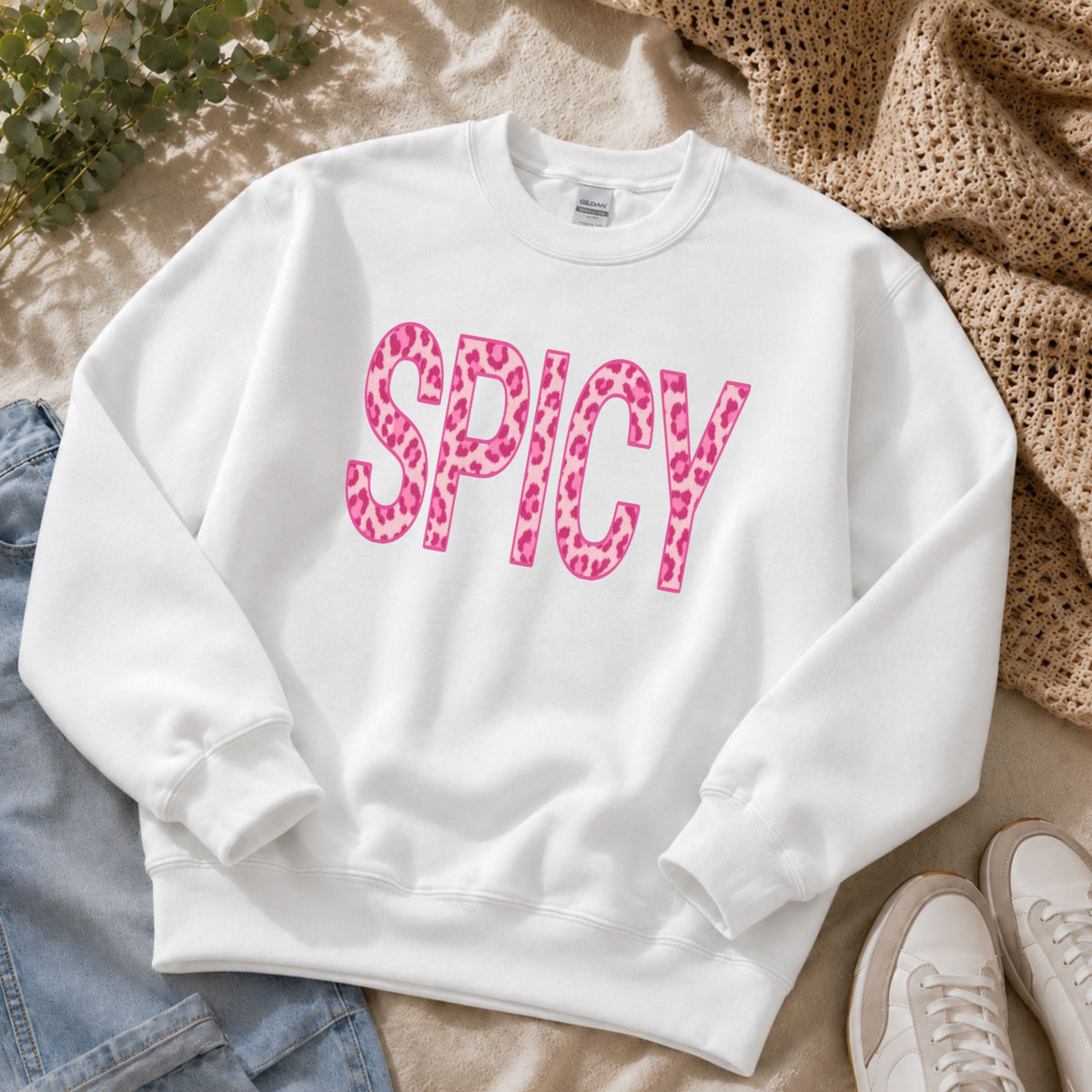 A white crewneck sweatshirt laid flat on a beige fabric surface, featuring the word 'SPICY' in large pink leopard-print letters across the chest. Jeans, a beige knit blanket, and white sneakers are partially visible around it as styling props.
