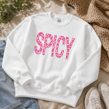 A white crewneck sweatshirt laid flat on a beige fabric surface, featuring the word 'SPICY' in large pink leopard-print letters across the chest. Jeans, a beige knit blanket, and white sneakers are partially visible around it as styling props.
