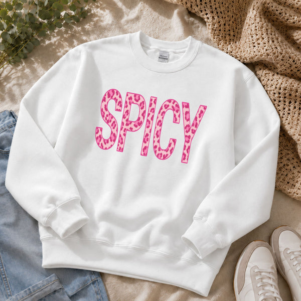 A white crewneck sweatshirt laid flat on a beige fabric surface, featuring the word 'SPICY' in large pink leopard-print letters across the chest. Jeans, a beige knit blanket, and white sneakers are partially visible around it as styling props.