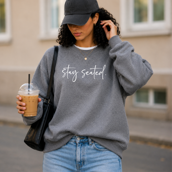 Woman wearing a gray sweatshirt with 'stay seated.' text, holding a drink, outdoors.