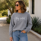 Woman wearing a gray sweatshirt with 'stay seated' text, standing outdoors.