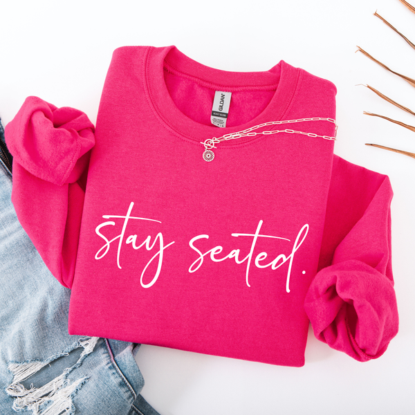 A heliconia bright pink folded sweatshirt on that says "Stay Seated" in a white script.