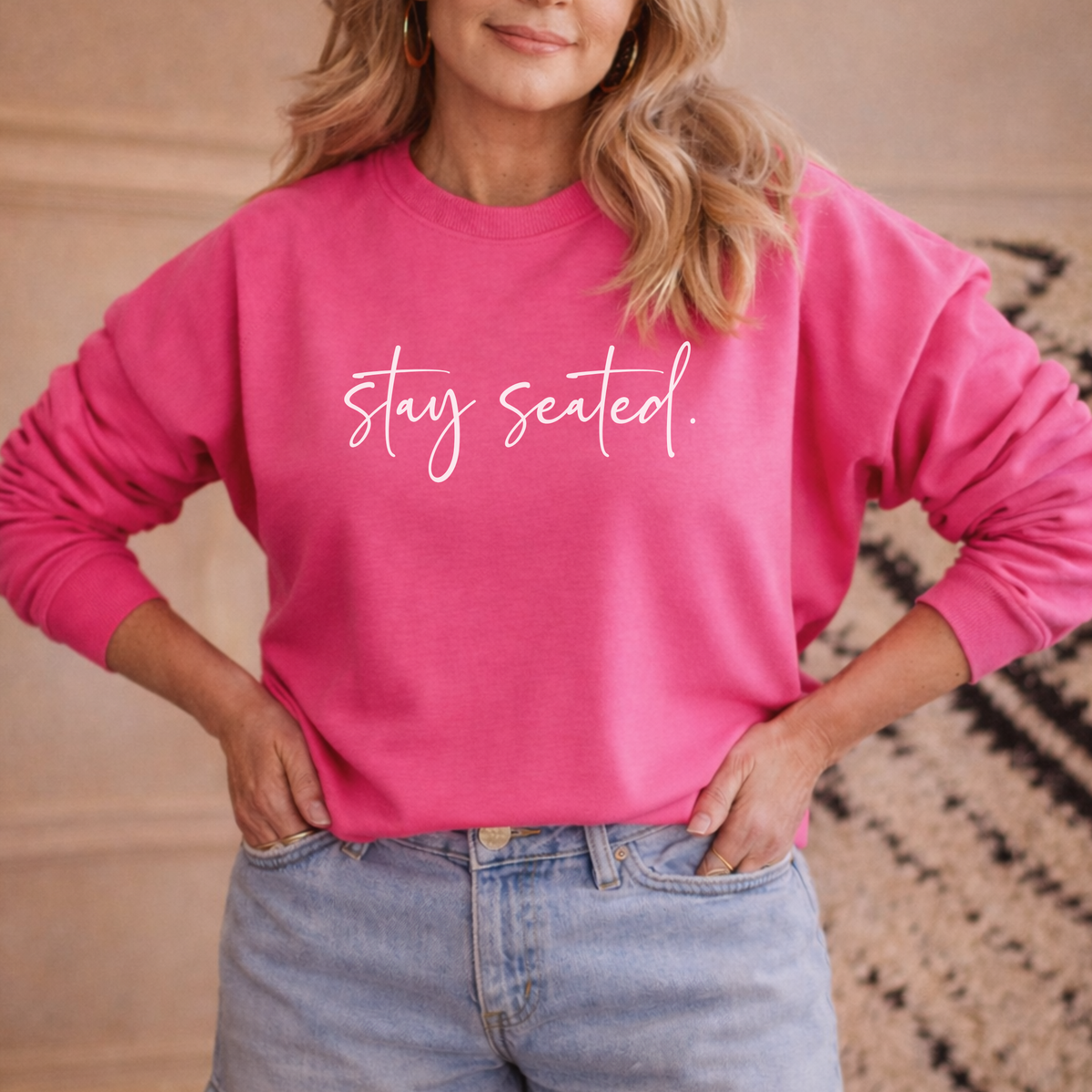 a 40-50 year old woman wearing a pink sweatshirt with 'stay seated' text, standing against a neutral background.