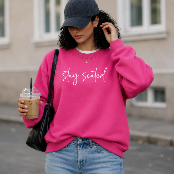 Woman wearing a pink sweatshirt with 'stay seated.' text, holding a drink, standing outdoors.