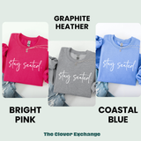 Three sweatshirts in different colors with 'stay seated.' text, labeled Graphite Heather, Bright Pink, and Coastal Blue, on a light gray background.