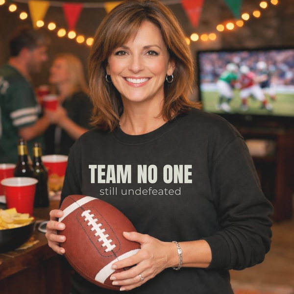 Woman wearing a black sweatshirt with 'TEAM NO ONE still undefeated' holding a football in a festive setting.