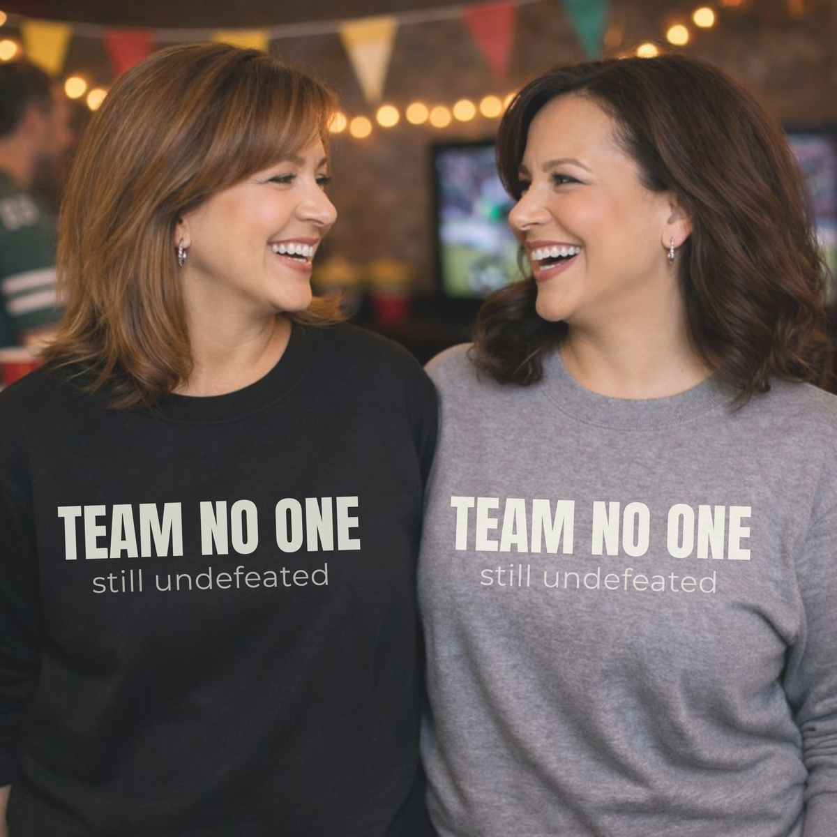 Two women wearing sweatshirts with 'TEAM NO ONE still undefeated' text, smiling and standing close together at a sports party.