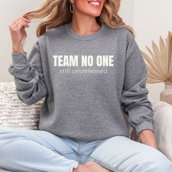 Person wearing a gray sweatshirt with 'TEAM NO ONE still undefeated' text, sitting on a couch.