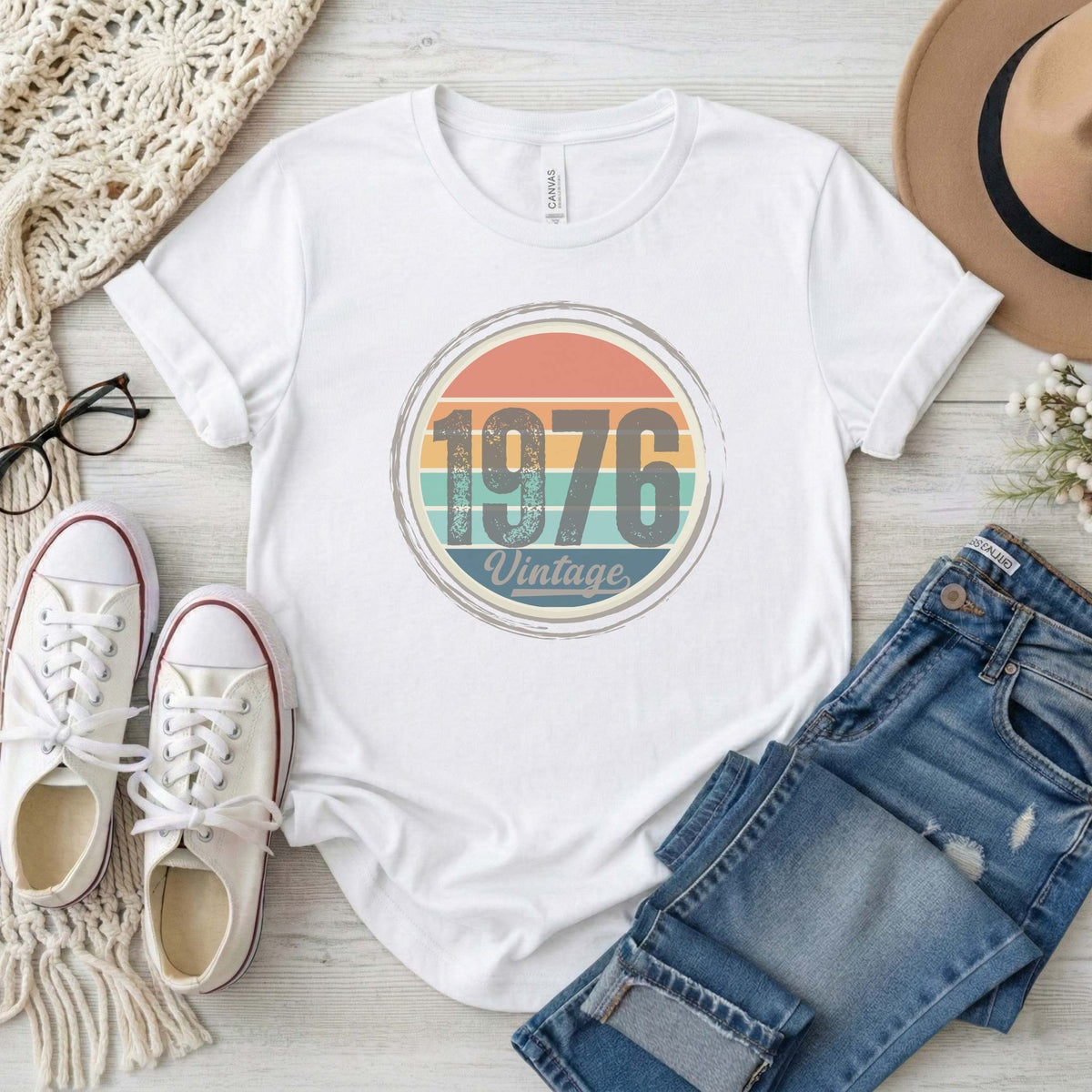 White t-shirt with '1976 Vintage' design, paired with jeans, sneakers, and a hat on a light wooden surface.