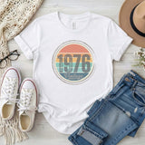 White t-shirt with '1976 Vintage' design, paired with jeans, sneakers, and a hat on a light wooden surface.