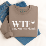 Folded brown sweatshirt with 'WTF wine turkey family' text and a wine glass graphic on blue jeans.