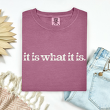 Mauve t-shirt with 'it is what it is' text on a white background with decorative elements.