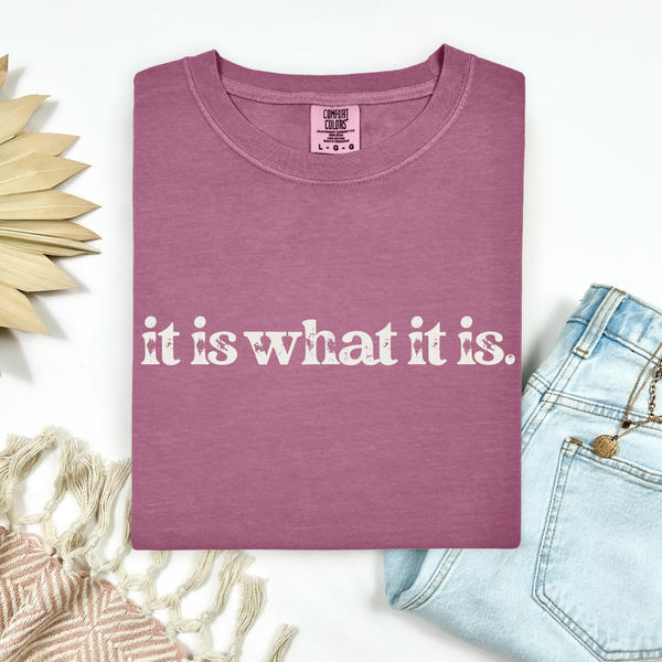 Mauve t-shirt with 'it is what it is' text on a white background with decorative elements.