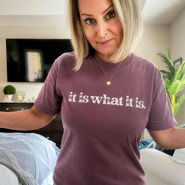 A woman wearing a heathered mauve short-sleeve t-shirt with the phrase 'it is what it is.' printed in white across the chest, standing in a bedroom setting.