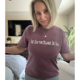 Woman wearing a purple t-shirt with 'it is what it is' text in a bedroom setting.