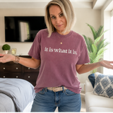 A woman wearing a mauve-purple graphic t-shirt with the white text 'it is what it is.' and blue jeans, standing in a brightly lit bedroom.