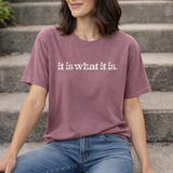 Person wearing a purple t-shirt with 'it is what it is' text sitting on steps.