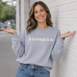 Woman wearing a gray sweatshirt with 'it is what it is' text, standing against a light-colored wall.