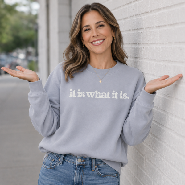 Woman wearing a gray sweatshirt with 'it is what it is' text, standing against a light-colored wall.