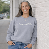 Woman wearing a gray sweatshirt with 'it is what it is' text, leaning against a light-colored wall.