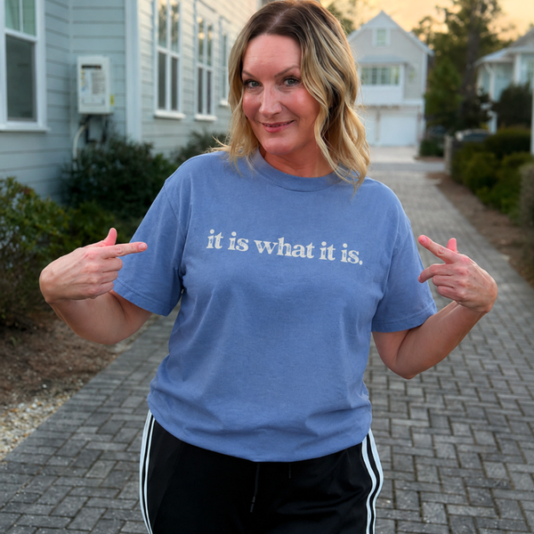 Person wearing a blue t-shirt with 'it is what it is' text, standing outdoors.