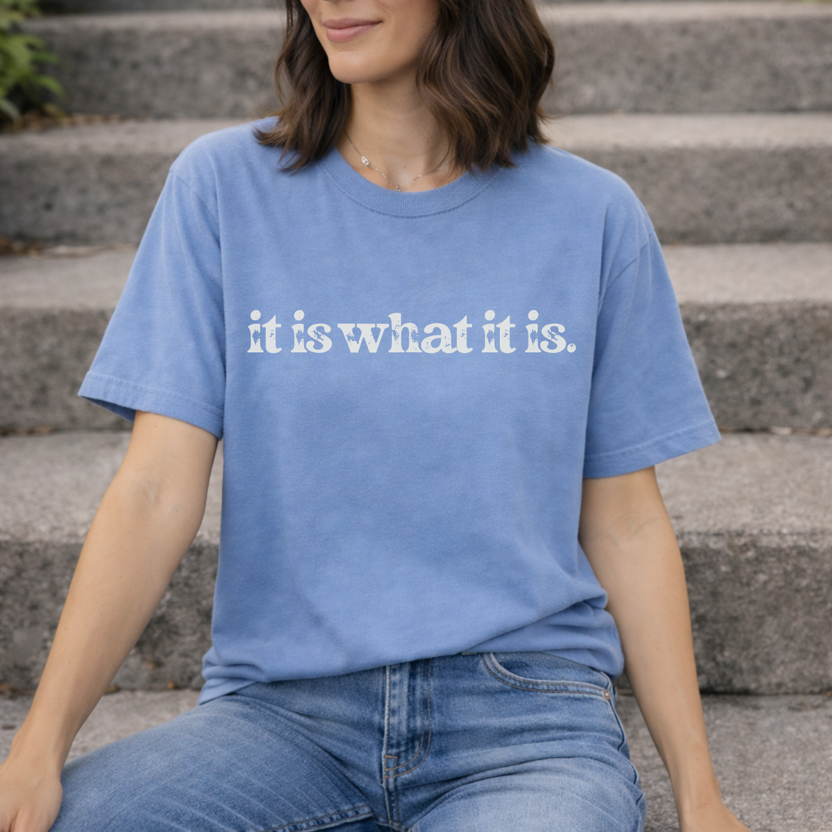 Person wearing a blue t-shirt with 'it is what it is' text sitting on steps.