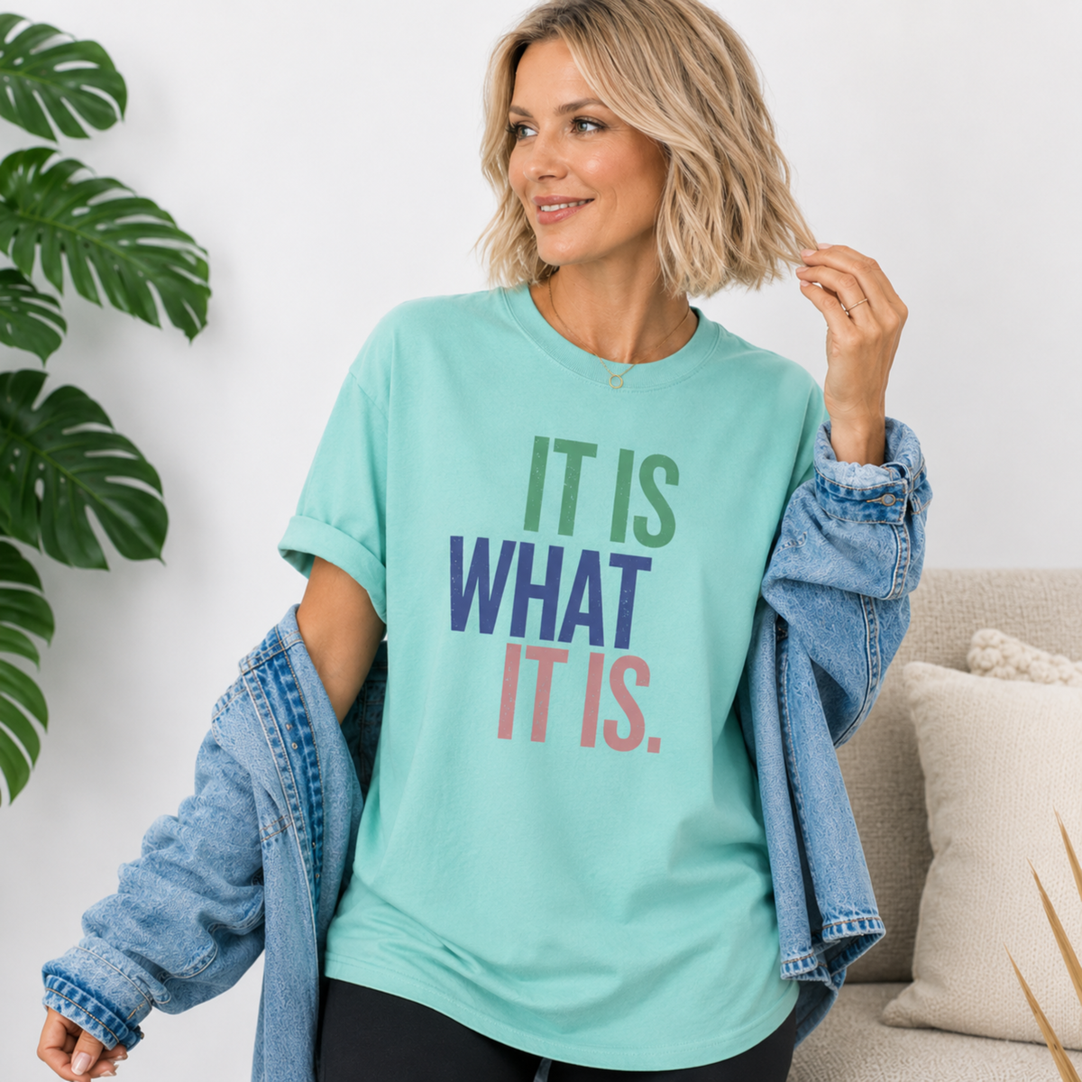 Woman wearing a light blue t-shirt with motivational text, standing indoors with a plant and couch in the background.