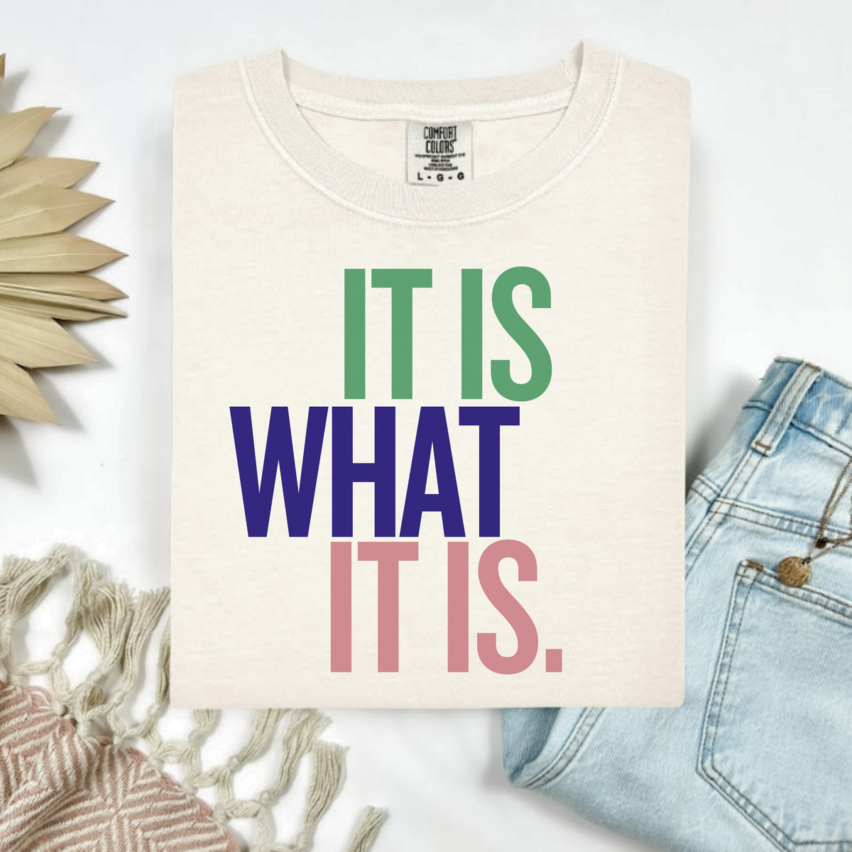 Beige t-shirt with colorful text 'IT IS WHAT IT IS' on a light background with denim jeans and decorative elements.
