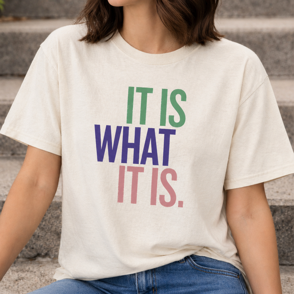 Person wearing a white t-shirt with colorful text 'IT IS WHAT IT IS' on a neutral background.