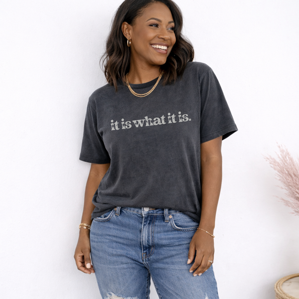 Woman wearing a dark gray t-shirt with 'it is what it is' text, standing against a white wall.