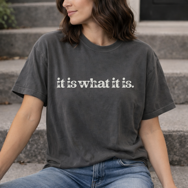 A woman wearing a dark gray t-shirt with 'it is what it is' text, sitting on steps.