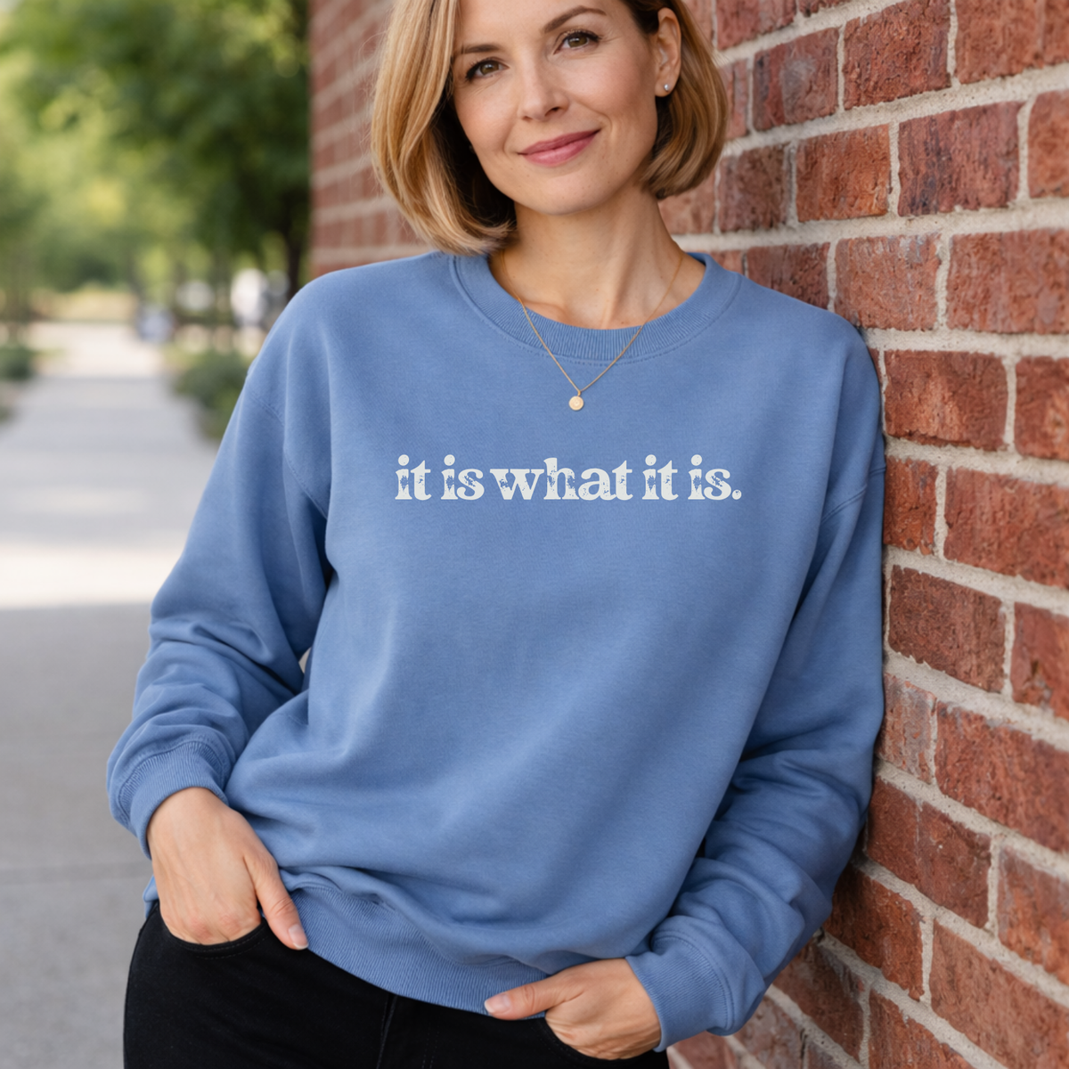 Woman wearing a blue sweatshirt with text that says "It is what it is" leaning against a brick wall.
