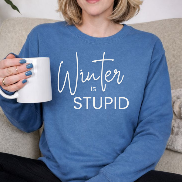 A woman  wearing a blue sweatshirt with 'Winter is STUPID' text, holding a white mug.