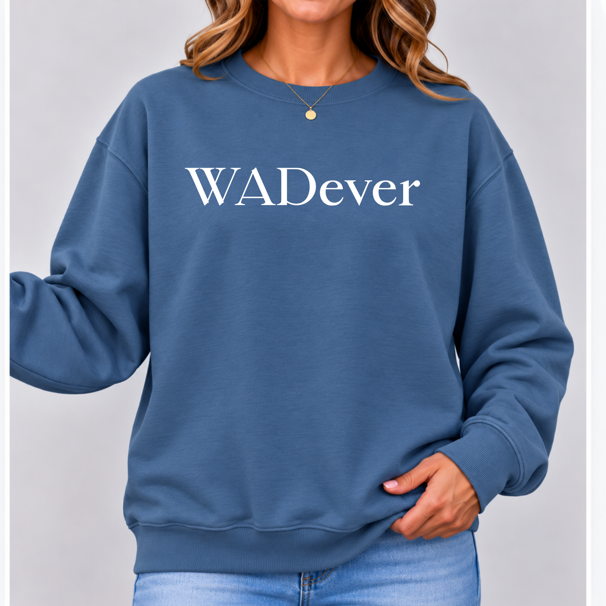 A woman wearing a slate blue crewneck sweatshirt with the text 'WADever' printed in white across the chest, paired with light blue jeans against a neutral gray background.