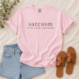 A light pink short-sleeve graphic T-shirt laid flat on a cream fabric background, styled with light blue denim shorts, brown slide sandals, and a small green leafy sprig. The shirt has dark olive text reading 'sarcasm IS MY LOVE LANGUAGE' across the chest.