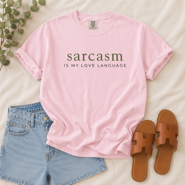 A light pink short-sleeve graphic T-shirt laid flat on a cream fabric background, styled with light blue denim shorts, brown slide sandals, and a small green leafy sprig. The shirt has dark olive text reading 'sarcasm IS MY LOVE LANGUAGE' across the chest.