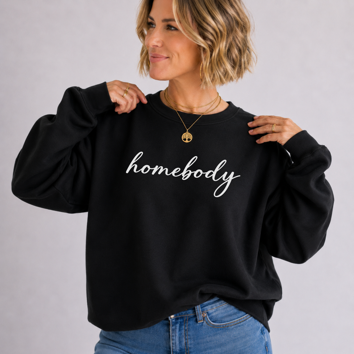 A woman wearing a black crewneck sweatshirt with the word 'homebody' printed in white script across the chest, paired with blue jeans, against a light gray studio background.