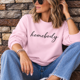 A woman wearing a light pink crewneck sweatshirt with the word 'homebody' printed in black script across the chest, paired with blue jeans and white sneakers, sitting against a stone wall.