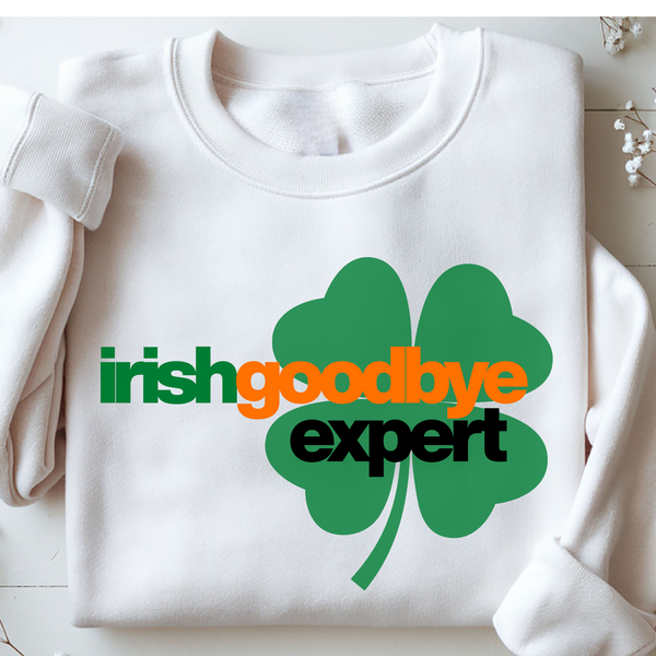 White sweatshirt with a green four-leaf clover and text 'irish goodbye expert' on a light wooden surface.