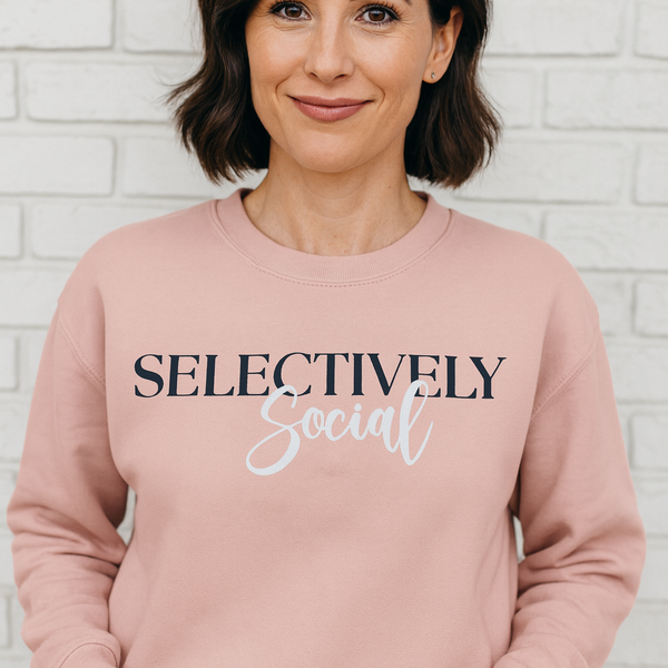 Woman wearing a pink sweatshirt with 'SELECTIVELY Social' text against a white brick wall.